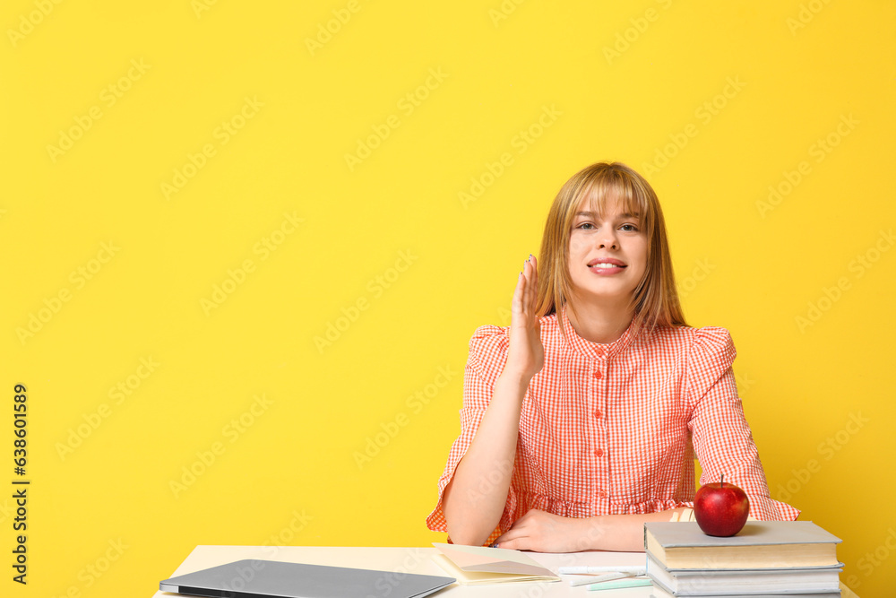 Female student raising hand at table on yellow background