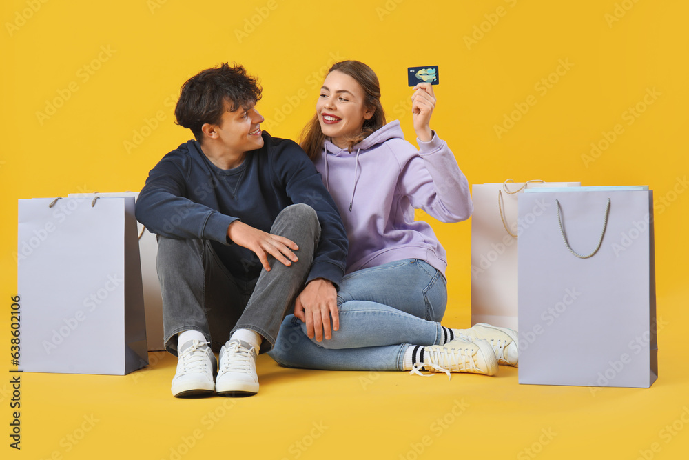 Happy young couple with credit card and shopping bags sitting on yellow background