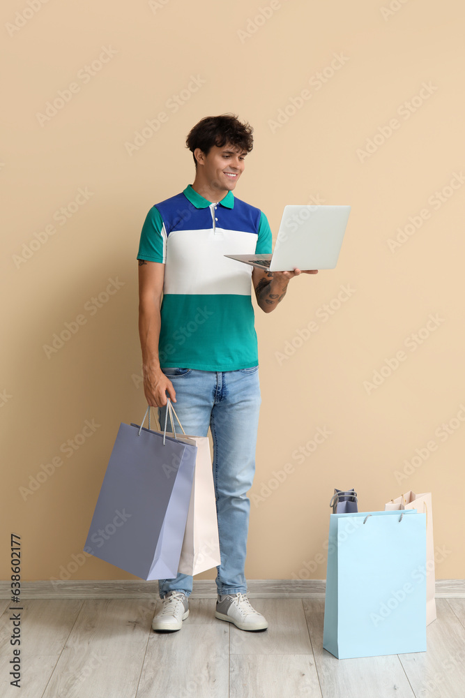 Young man with laptop and shopping bags near beige wall