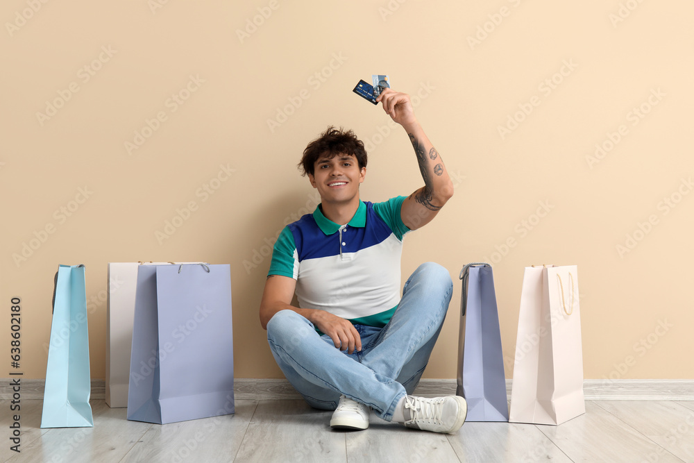 Young man with credit cards and shopping bags sitting near beige wall