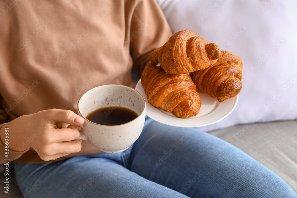 Young woman with tasty croissants and cup of coffee sitting on sofa in living room