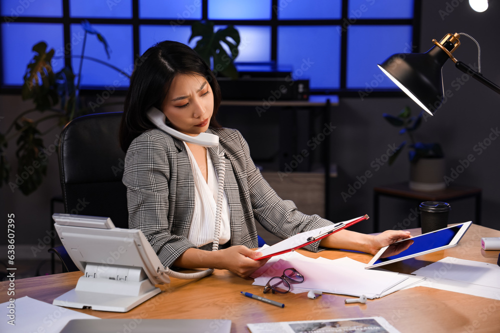 Young Asian businesswoman sitting at table, talking by phone and working in office