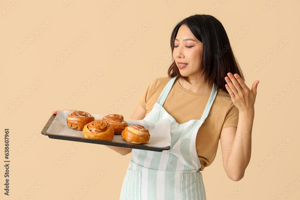 Happy beautiful Asian woman holding tray with fresh cinnamon rolls on beige background