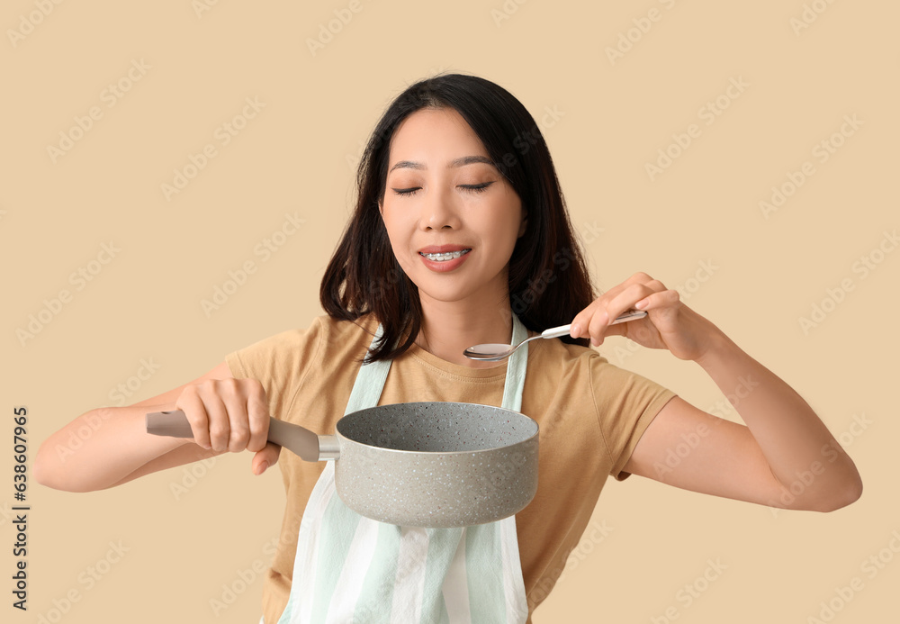 Happy beautiful Asian woman with saucepan and spoon on beige background