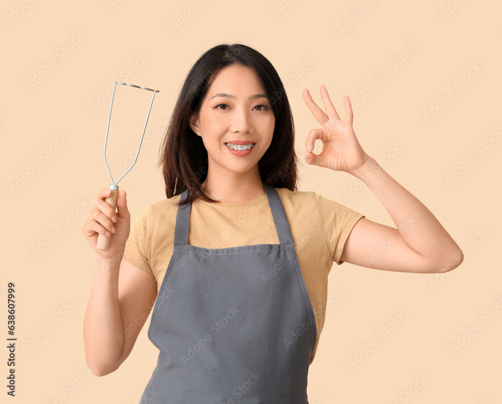 Happy beautiful Asian woman with masher showing OK gesture on beige background