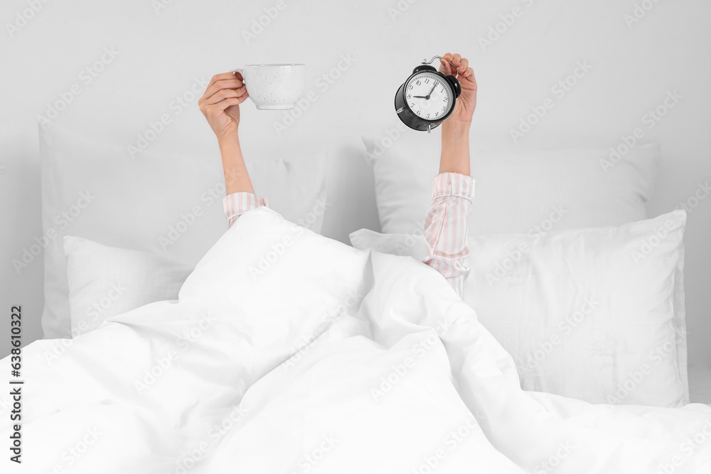 Woman holding cup of coffee and alarm clock on bed in bedroom