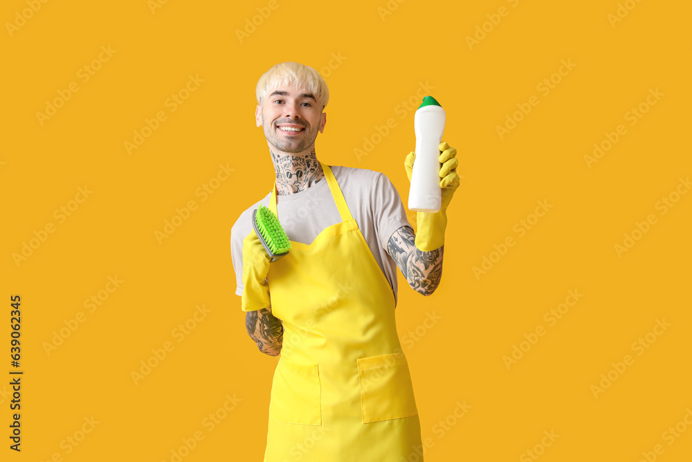 Young tattooed man with bottle of detergent and cleaning brush on orange background