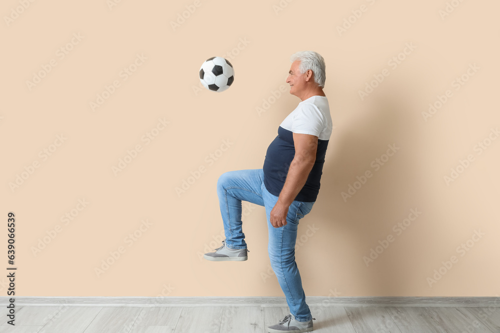 Mature man playing with soccer ball near beige wall