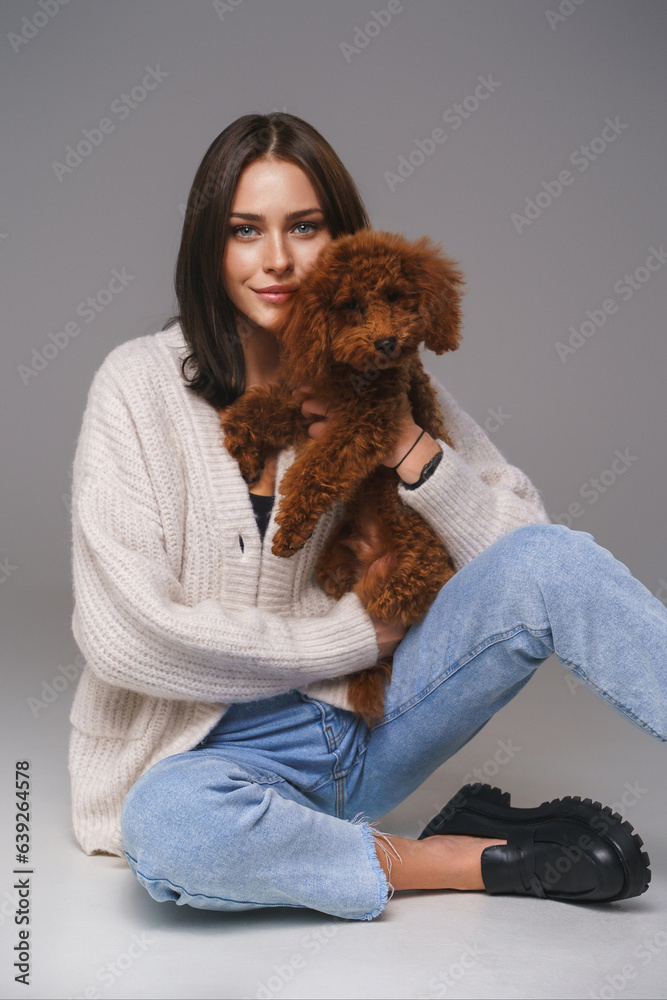 Elegant brunette in white top and denim shorts sits on the floor, adoringly holding her toy poodle, 