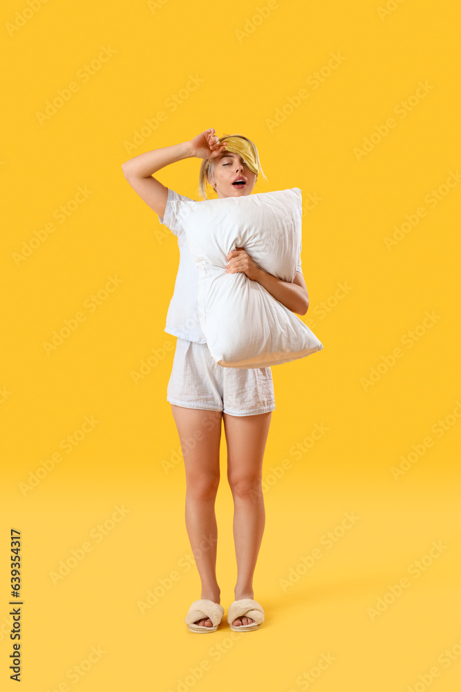 Young woman with pillow yawning on yellow background