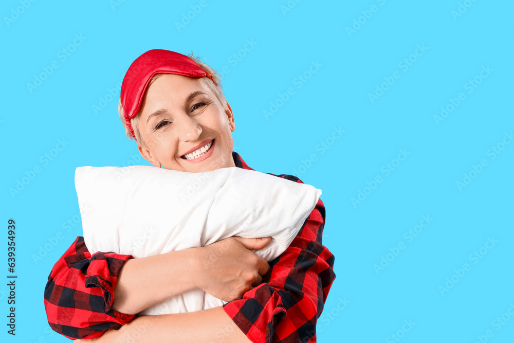 Mature woman in pajamas with pillow on blue background