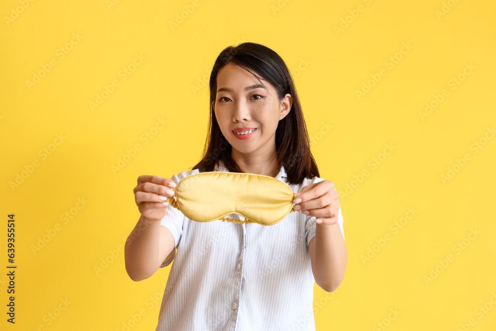 Beautiful Asian woman in pajamas with sleeping mask on yellow background