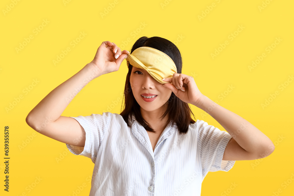 Beautiful Asian woman in pajamas with sleeping mask on yellow background