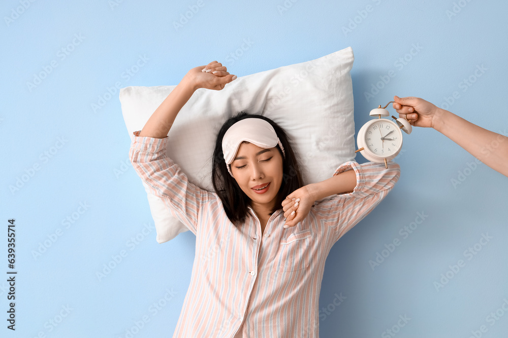 Beautiful Asian woman in pajamas with pillow and ringing alarm clock on blue background