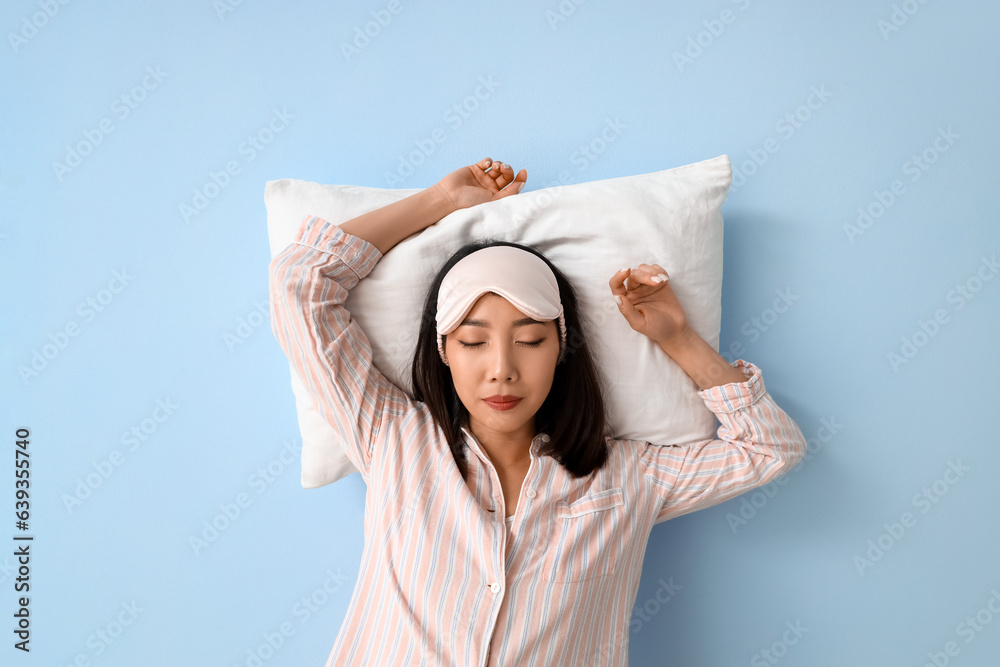 Beautiful Asian woman in pajamas with pillow sleeping on blue background