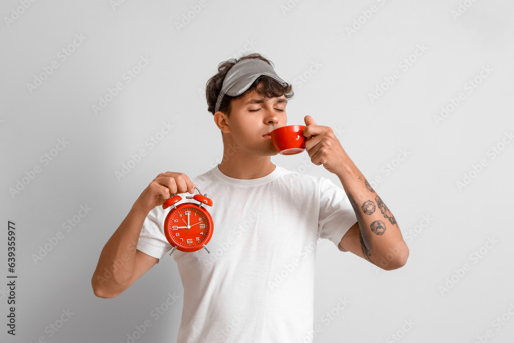 Young man with alarm clock drinking coffee on light background