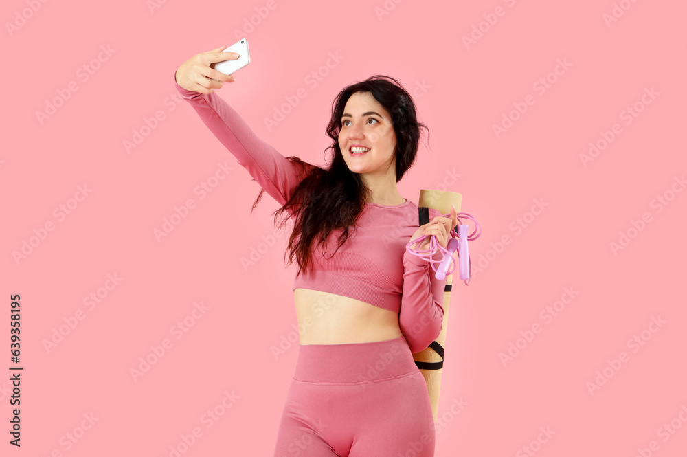 Sporty young woman with jumping rope and yoga mat taking selfie on pink background