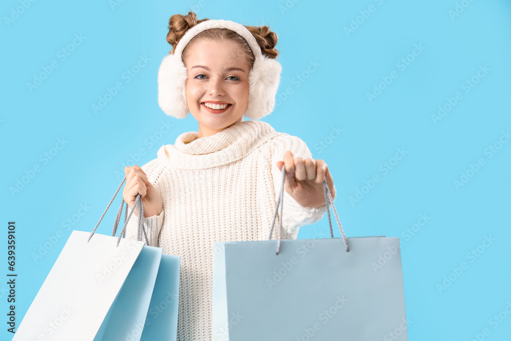 Happy young woman in warm sweater and fluffy headphones with shopping bags on blue background