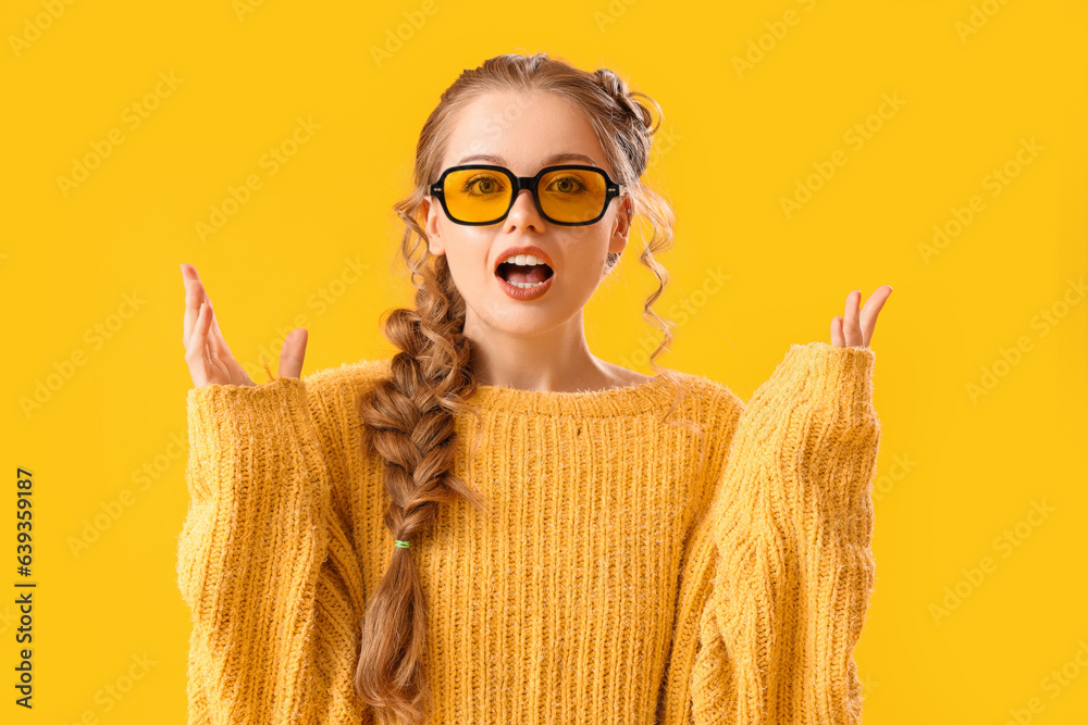 Beautiful shocked young woman in warm sweater and eyeglasses on yellow background