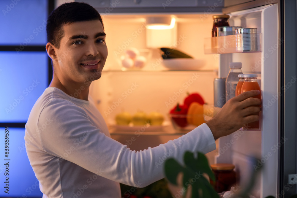 Hungry young man with bottle of juice near open fridge in kitchen at night