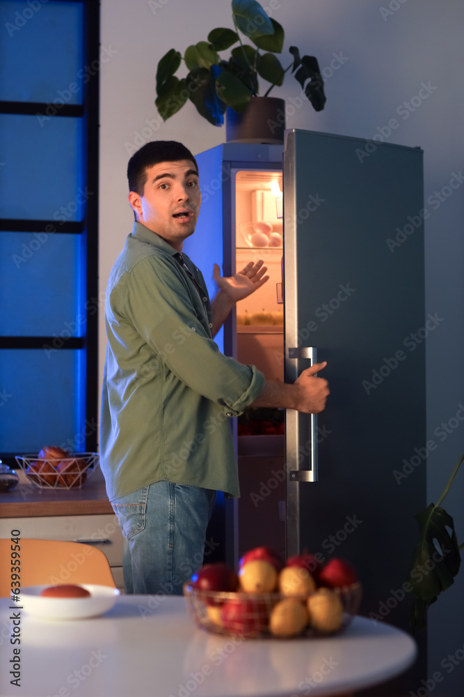 Hungry young man near open fridge in kitchen at night