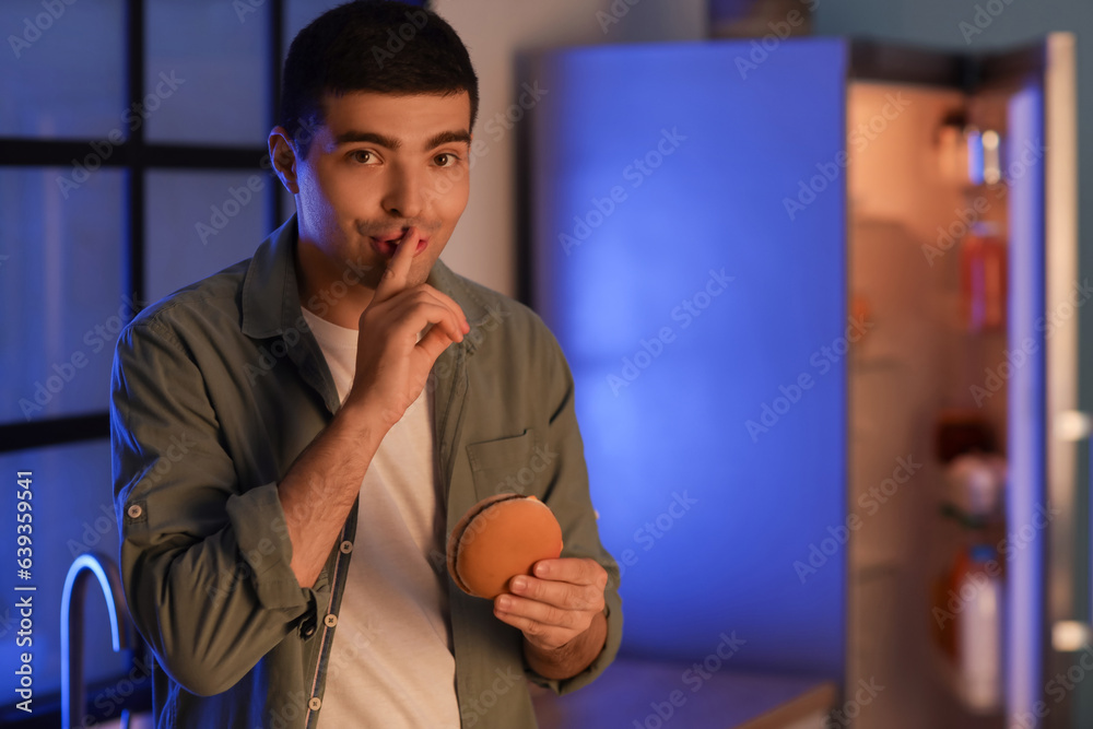 Hungry young man with burger showing silence gesture in kitchen at night