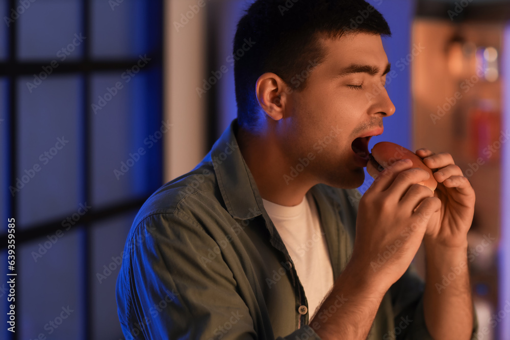 Hungry young man eating burger in kitchen at night, closeup