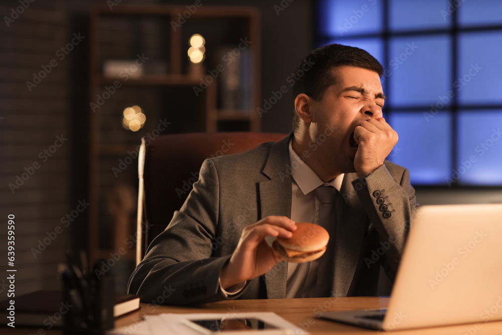 Sleepy young businessman with tasty burger in office at night