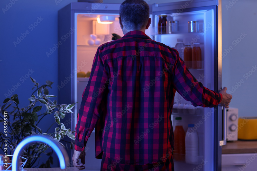 Hungry mature man near open fridge in kitchen at night, back view