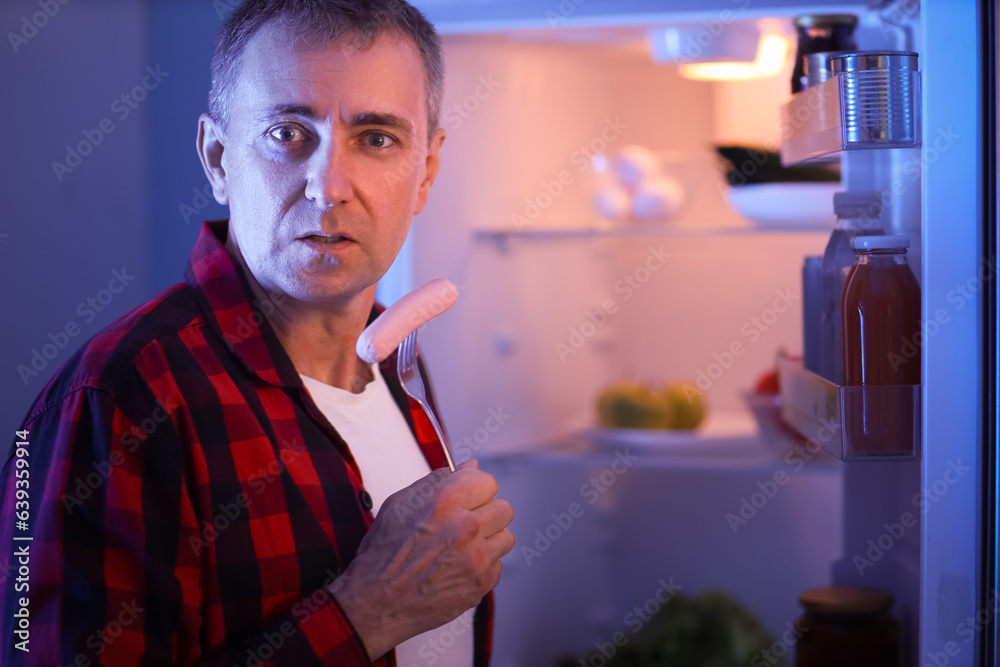 Hungry mature man with sausage near open fridge in kitchen at night, closeup