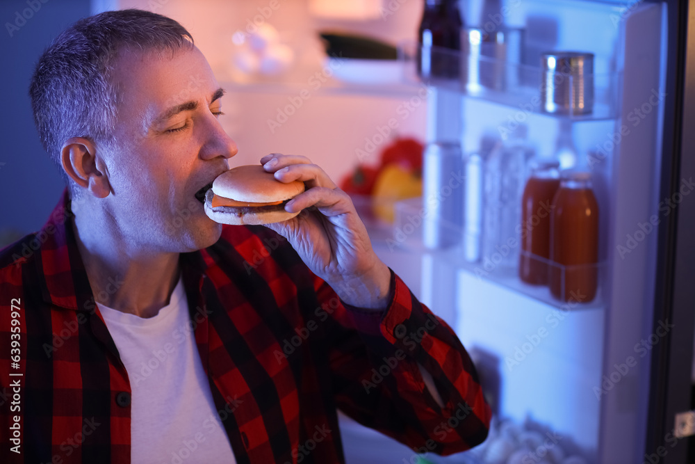 Hungry mature man eating burger near open fridge in kitchen at night, closeup