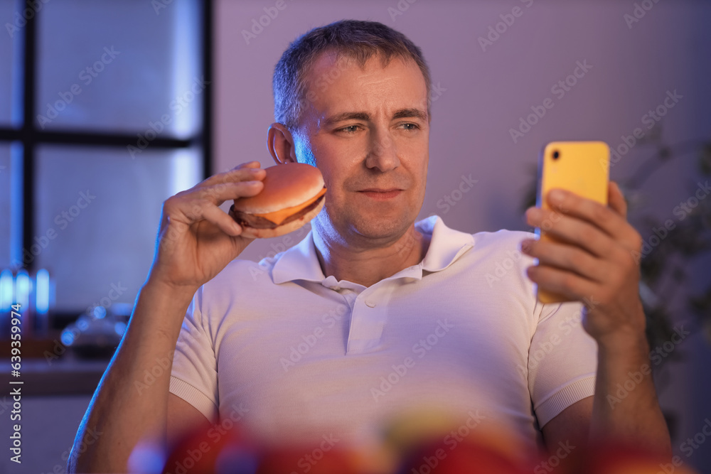 Hungry mature man with burger and mobile phone in kitchen at night