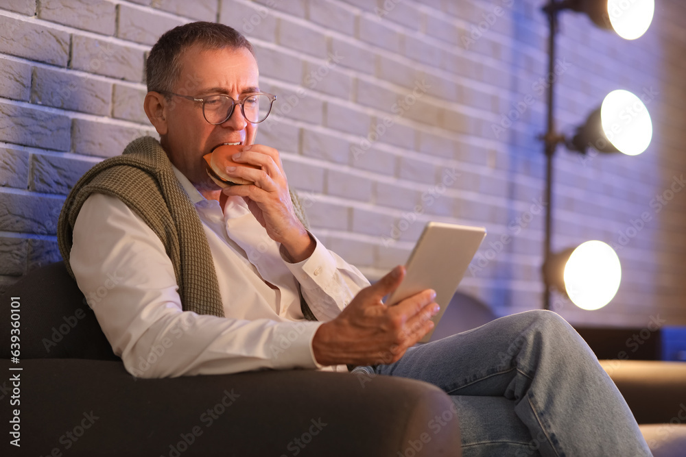 Mature man with tablet computer eating burger in office at night