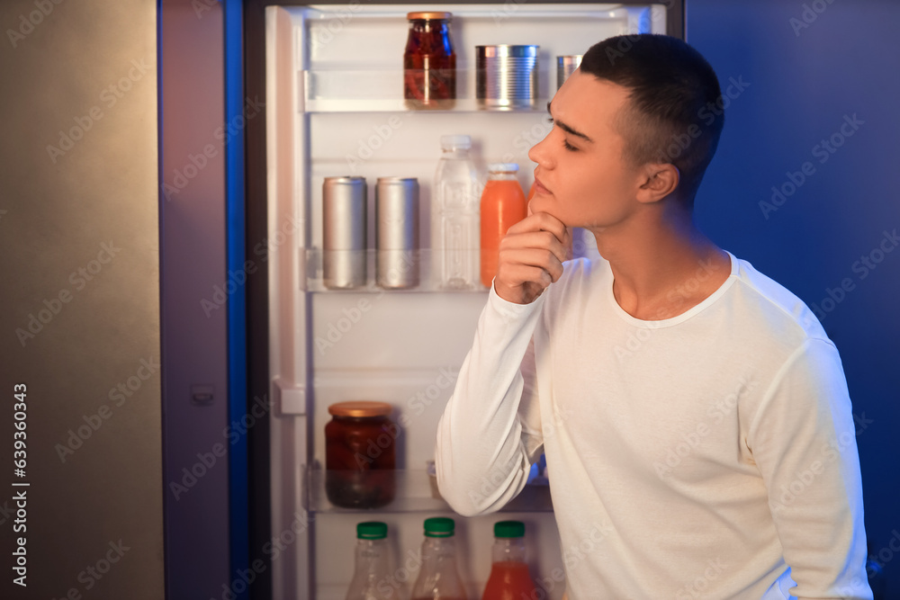 Hungry young man near open fridge in kitchen at night