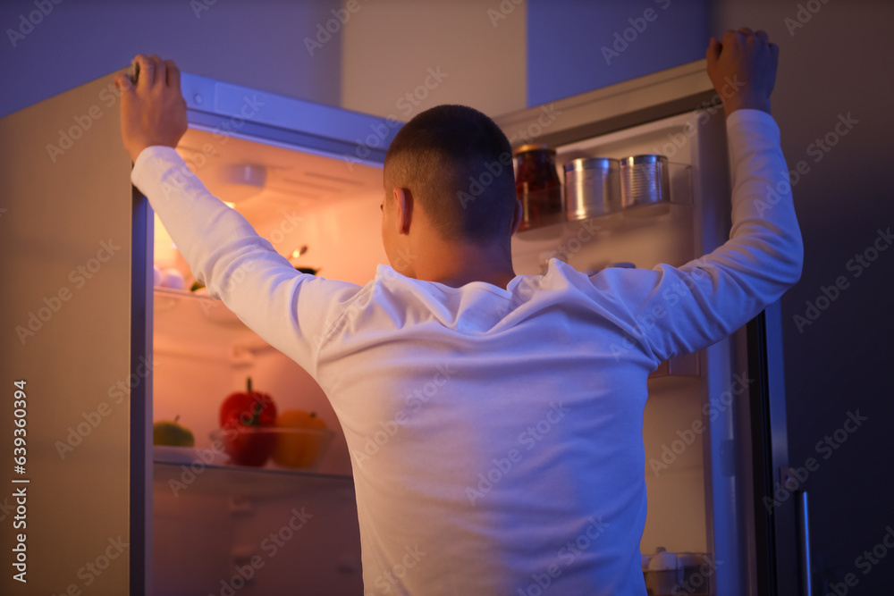 Hungry young man near open fridge in kitchen at night, back view