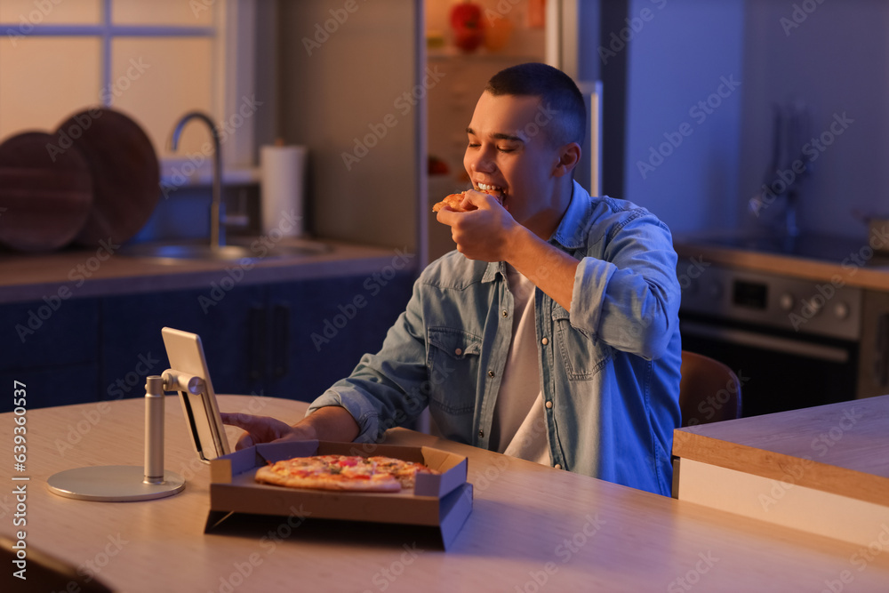 Hungry young man eating pizza in kitchen at night