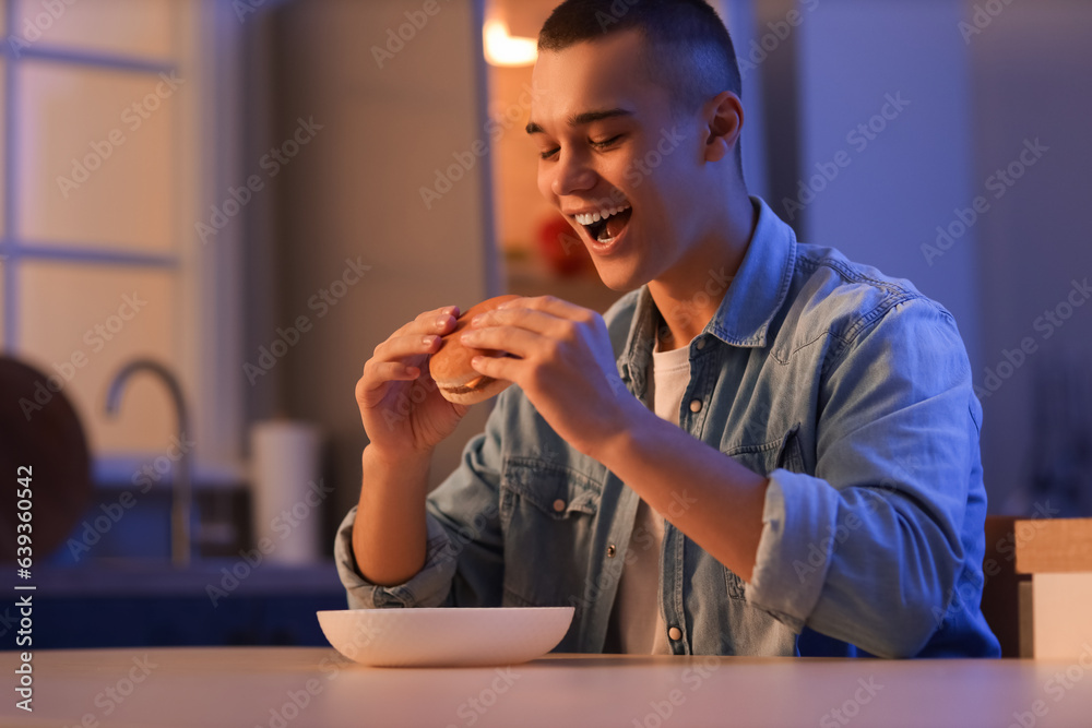 Hungry young man eating burger in kitchen at night