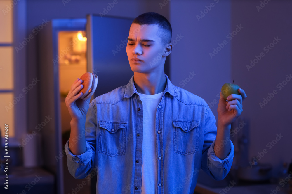 Thoughtful young man with burger and apple in kitchen at night