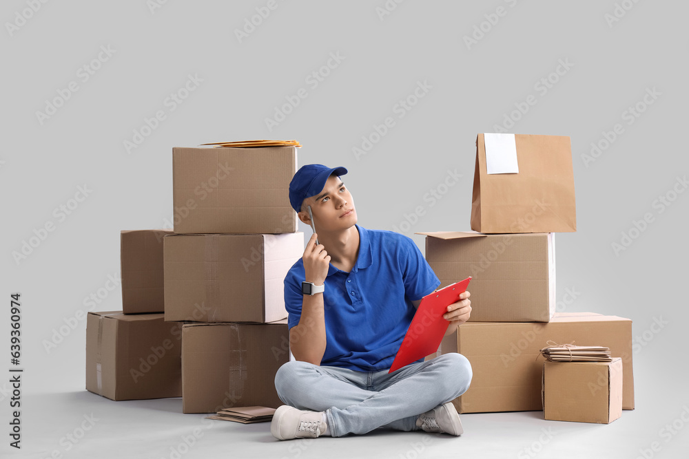 Thoughtful young courier with clipboard and parcels on light background