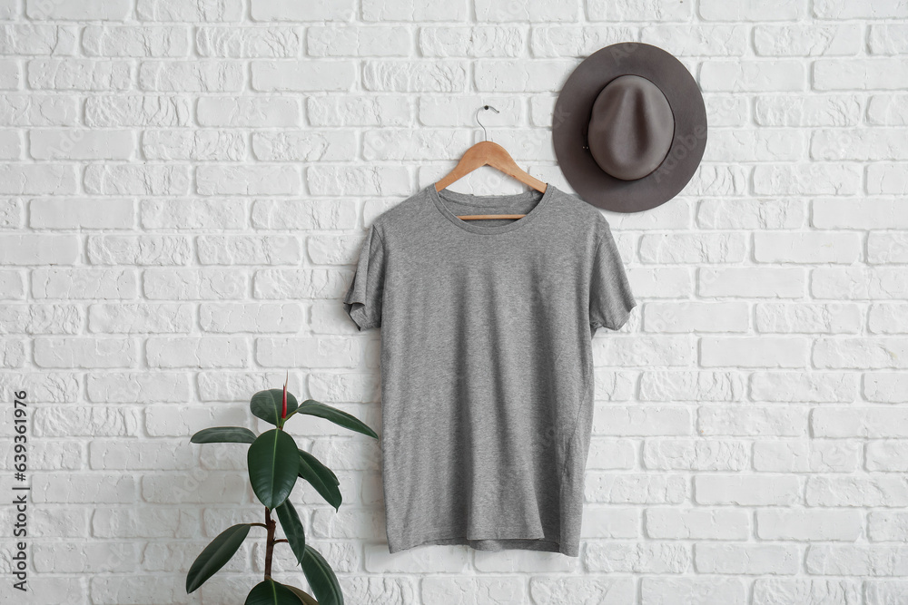 Stylish grey t-shirt and hat hanging on white brick wall