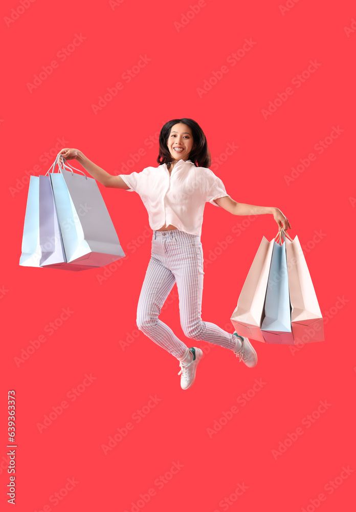 Beautiful stylish young Asian woman with shopping bags jumping on red background