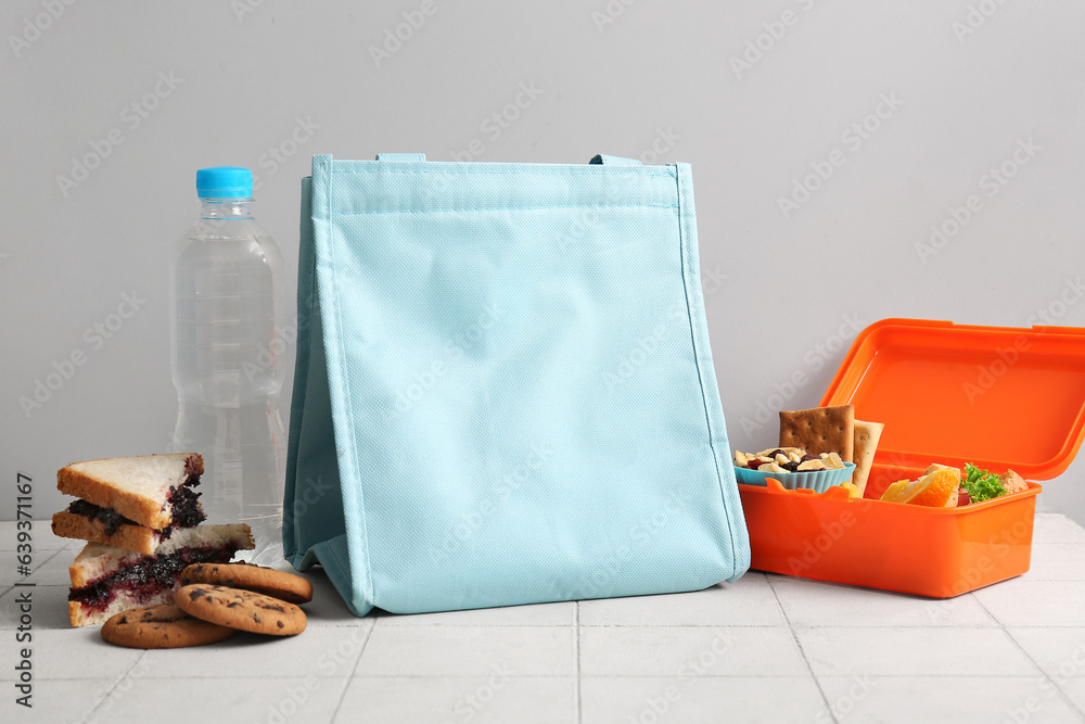 Bag, lunchbox with delicious food and bottle of water on white tile near grey wall