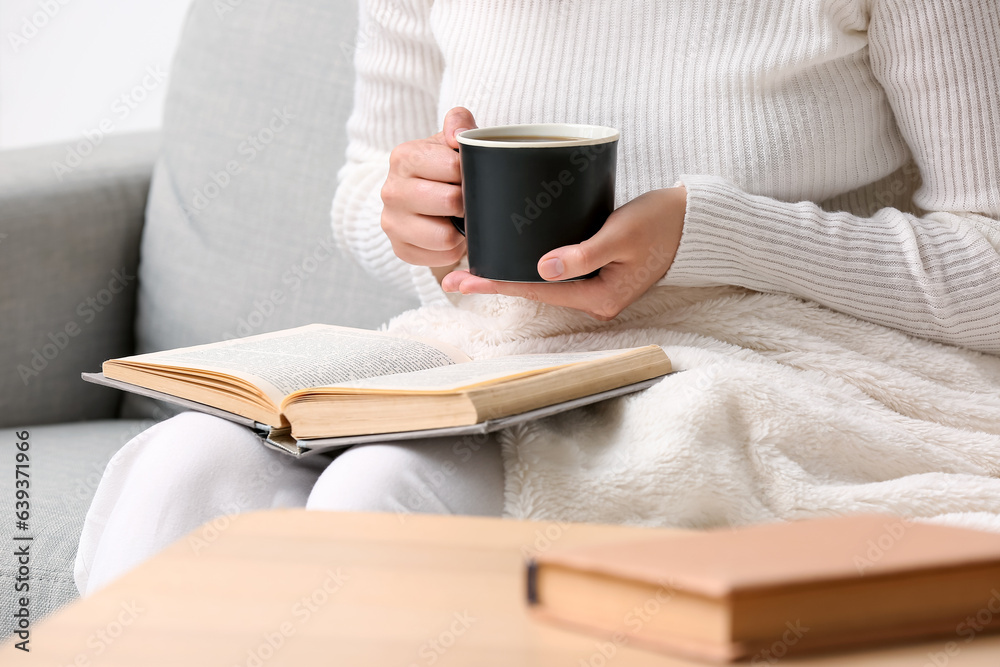 Woman with cup of tasty coffee and book resting on sofa at home