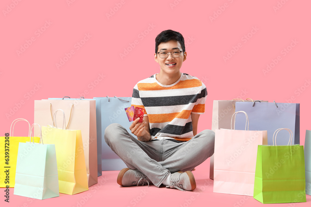 Young Asian man with gift cards and shopping bags sitting on pink background