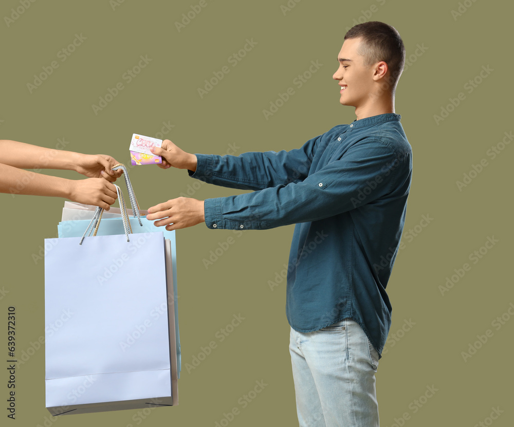 Young man with gift cards taking shopping bags on green background