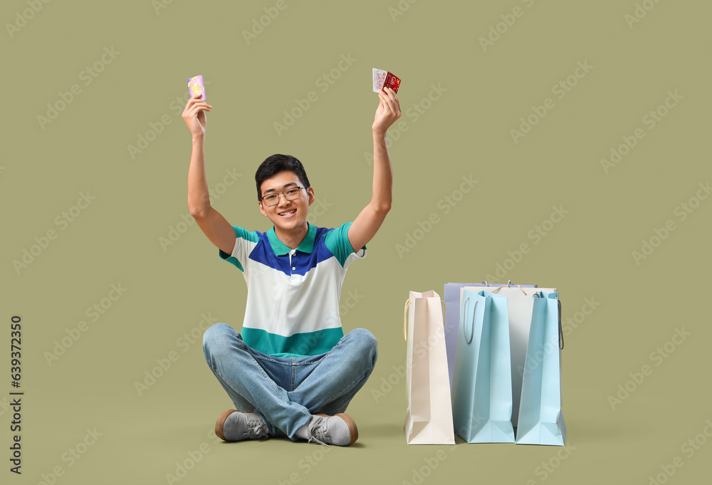 Young Asian man with gift cards and shopping bags on green background