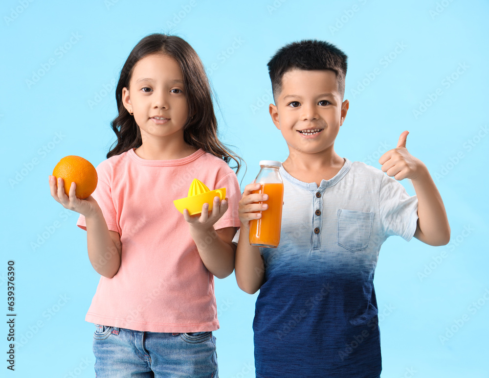 Little Asian children with fresh citrus juice, orange and squeezer showing thumb-up gesture on blue 