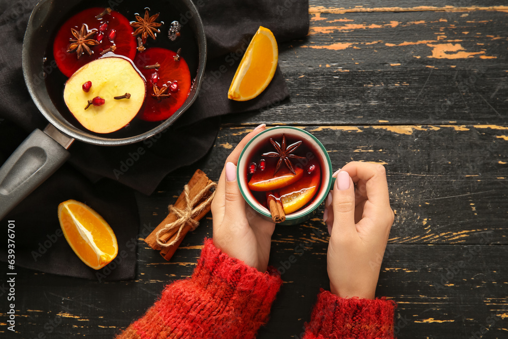 Woman holding cup of hot mulled wine with orange and cinnamon on black wooden background