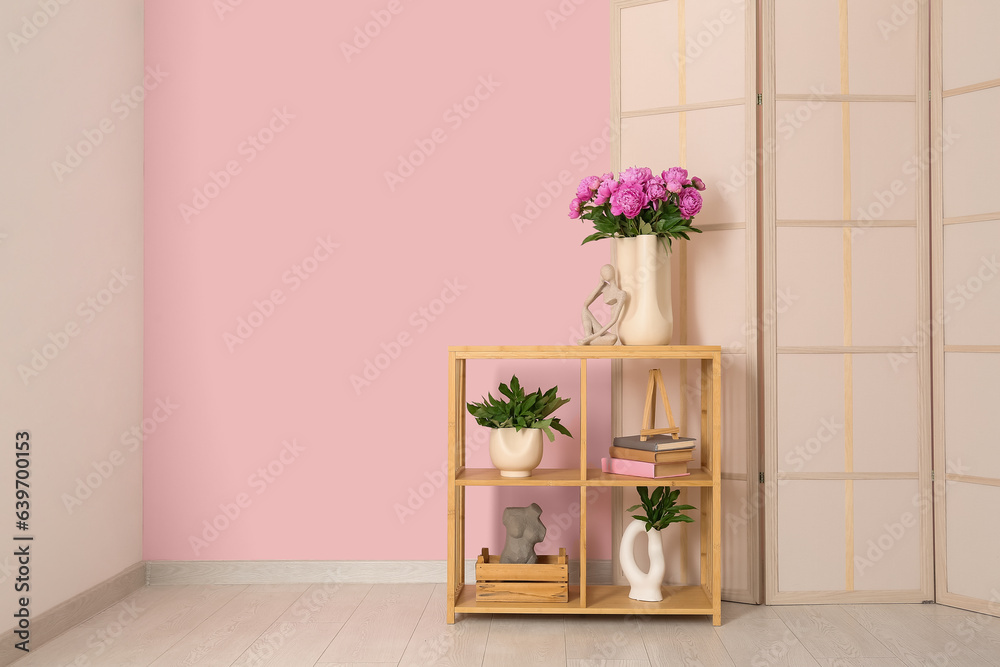 Vase of pink peonies and figurine on shelving unit with dressing screen near color wall