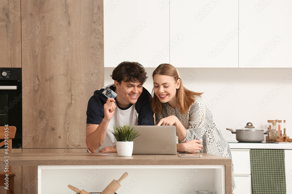 Young couple with laptop and gift card shopping online in kitchen